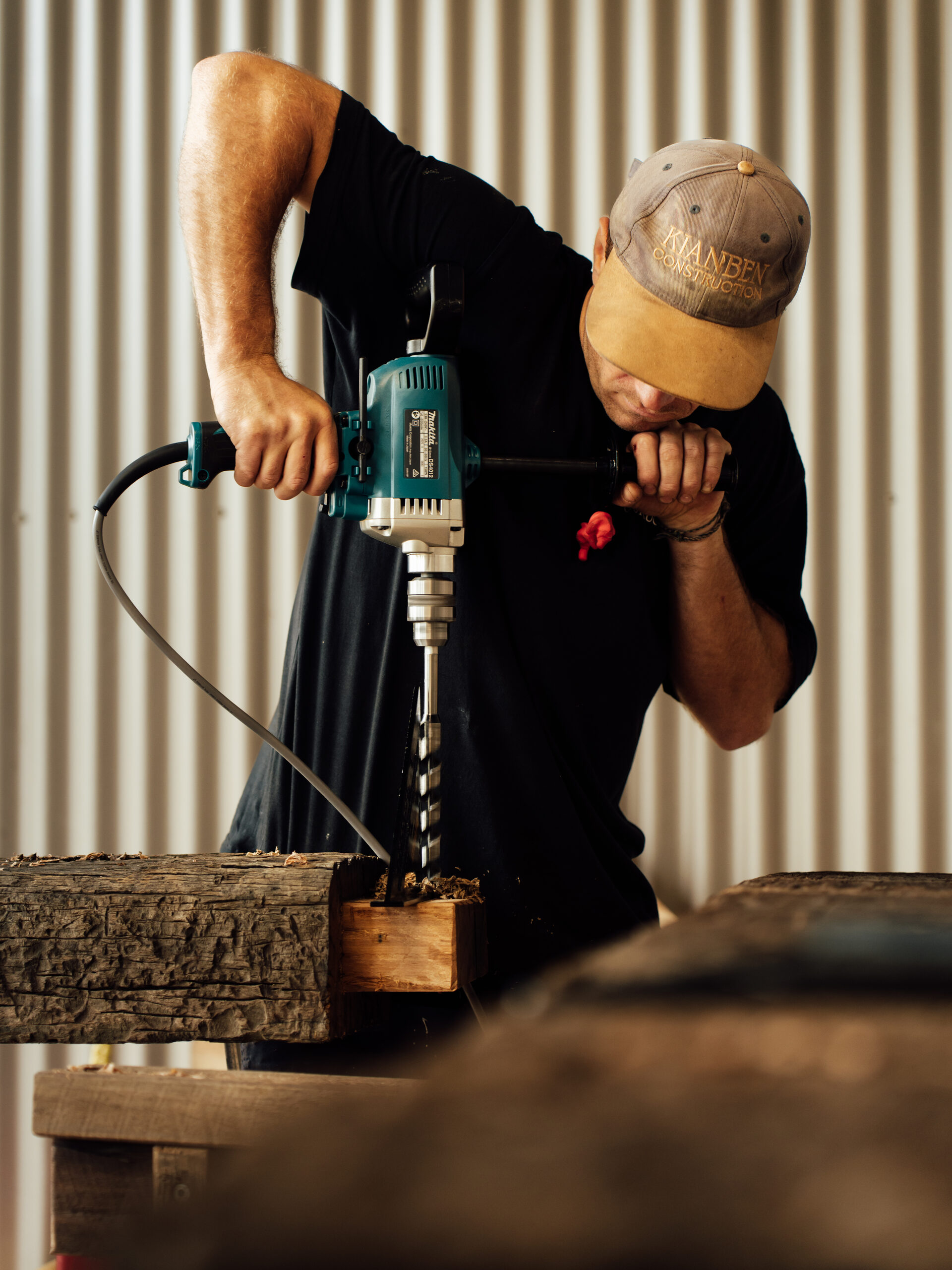 Boring a recycled hardwood beam with a drill, corrugated steel behind